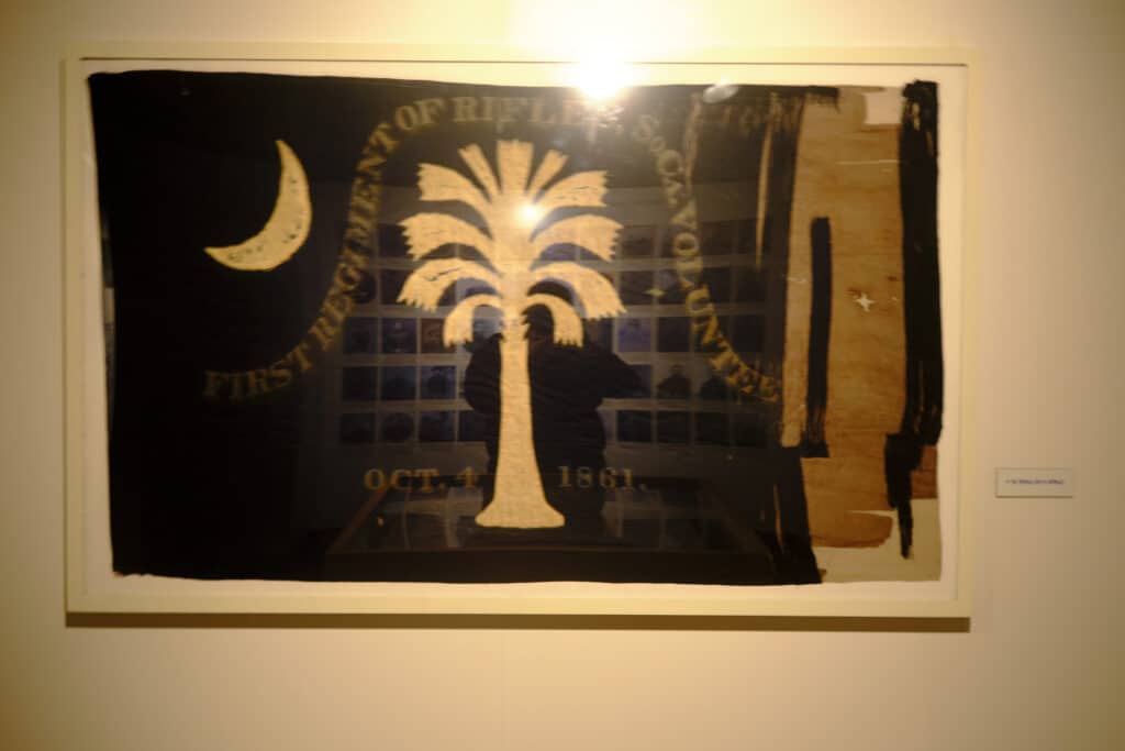 Photo of a flag on display at the SC Confederate Relic Room and Military Museum showing a flag that looks similar to the South Carolina state flag with palmetto tree and crescent.
