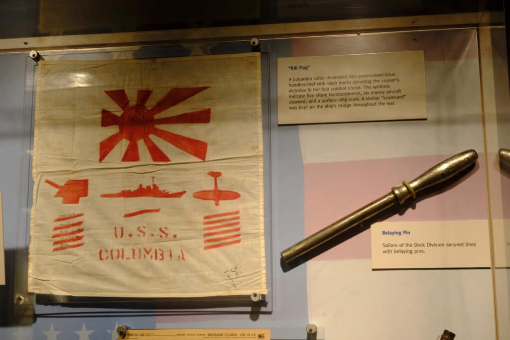 Photo of a museum display with a flag saying USS Columbia and showing lines below a Japanese flag next to another artifact.