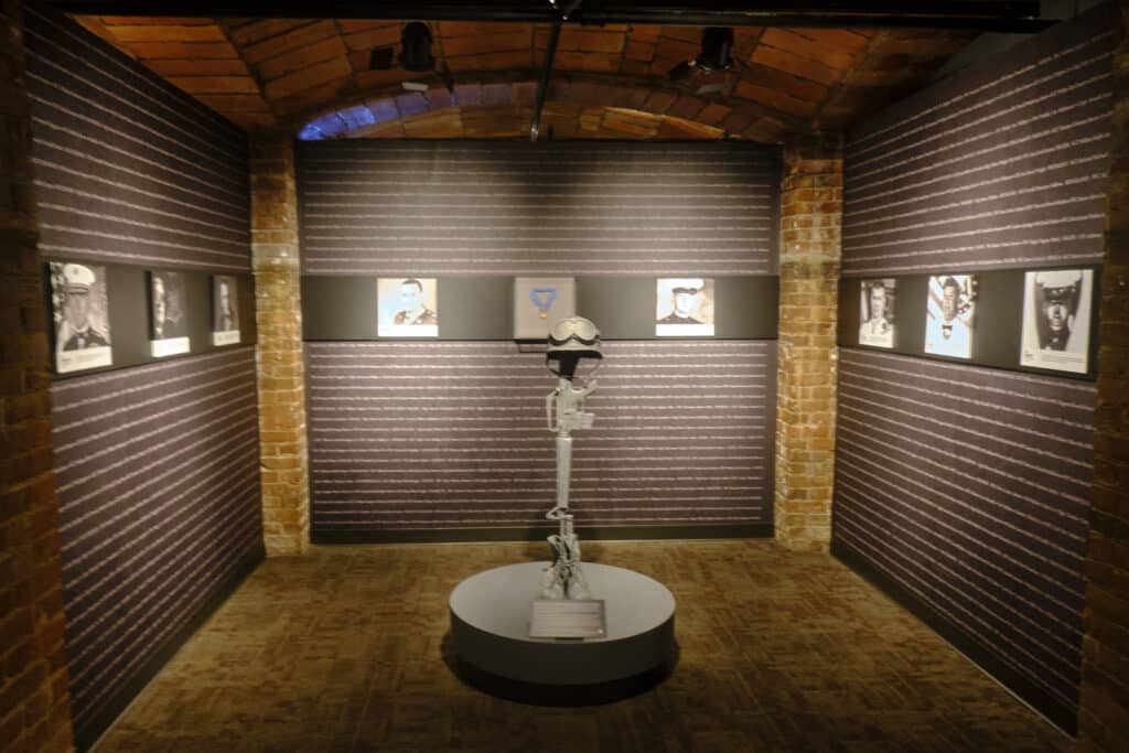 A memorial at the SC Confederate Relic Room and Military Museum showing a rifle with a helmet in a room with walls listing names and some photos.