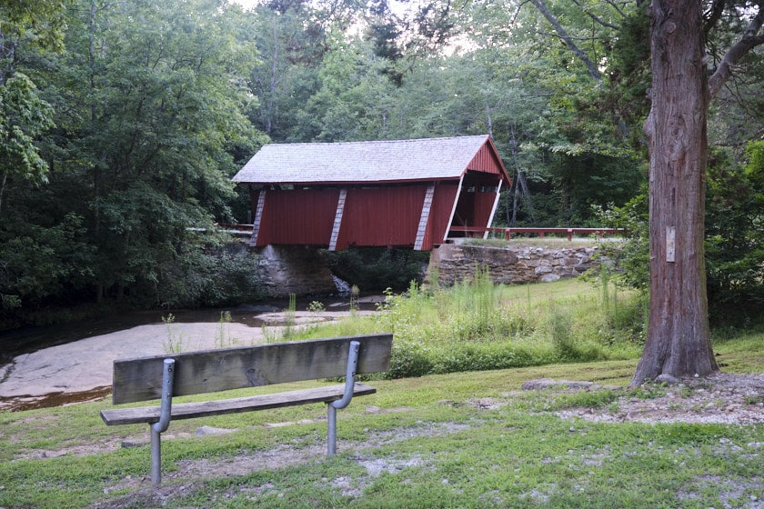 Photo of an empty bench overlooking a creek and the Campbell's Covered Bridge with a tree to the right.