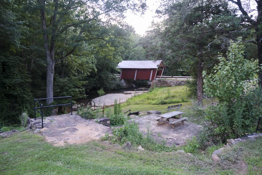 Photo of a creek with a picnic table and bench in the foreground and the red Campbell's Covered Bridge in the background through some trees.