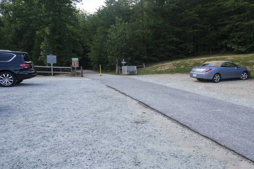 Photo of a gravel parking lot with paved road in the middle and two cars parked on either side of it, with a pathway leading into trees in the background.