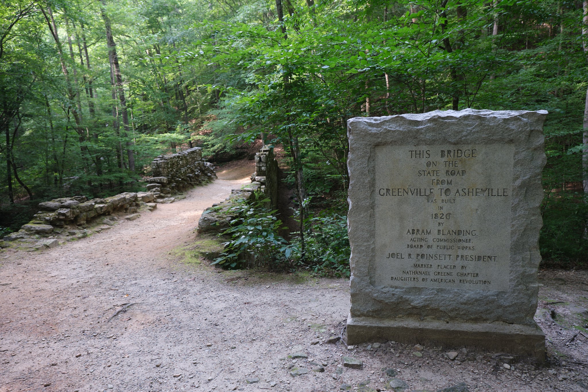 Photo of the Poinsett Bridge in South Carolina with a stone rectangle historic marker in the foreground and trees surrounding it.