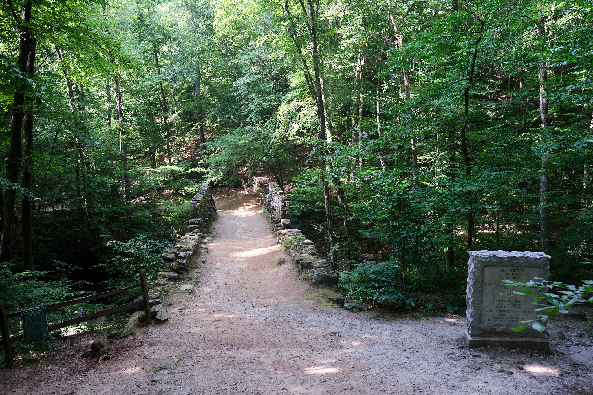 Photo of the Poinsett Bridge Heritage Preserve showing a pathway between two low stone walls and a rectangular stone marker to the left before the bridge.