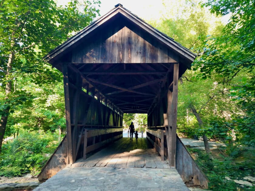 A woman and child holding hands crossing the Pisgah Covered Bridge from a distance.