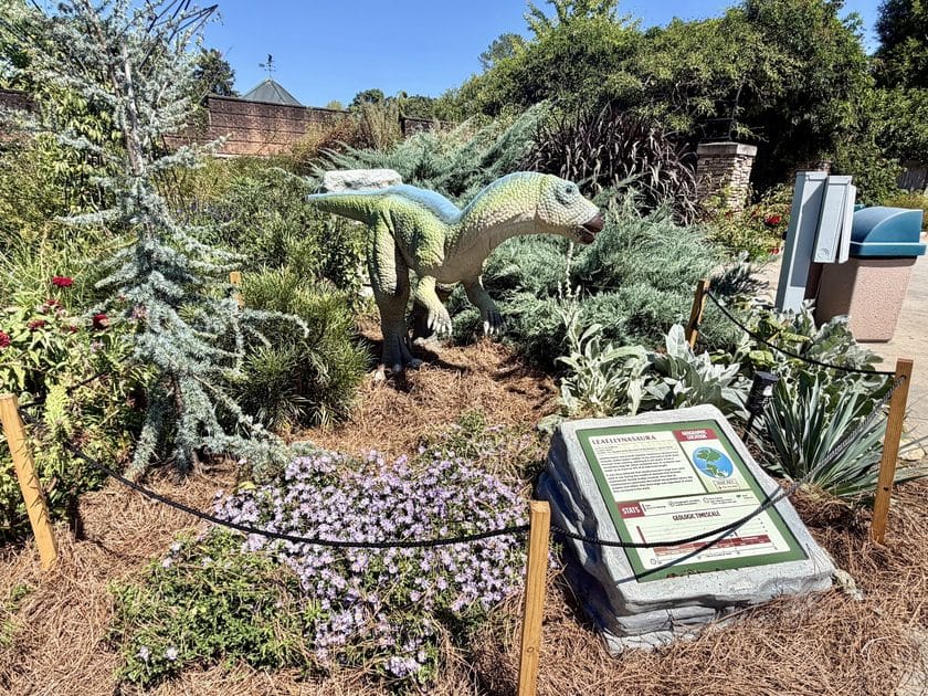 A small dinosaur at Riverbanks Zoo behind a sign and surrounded by plants.